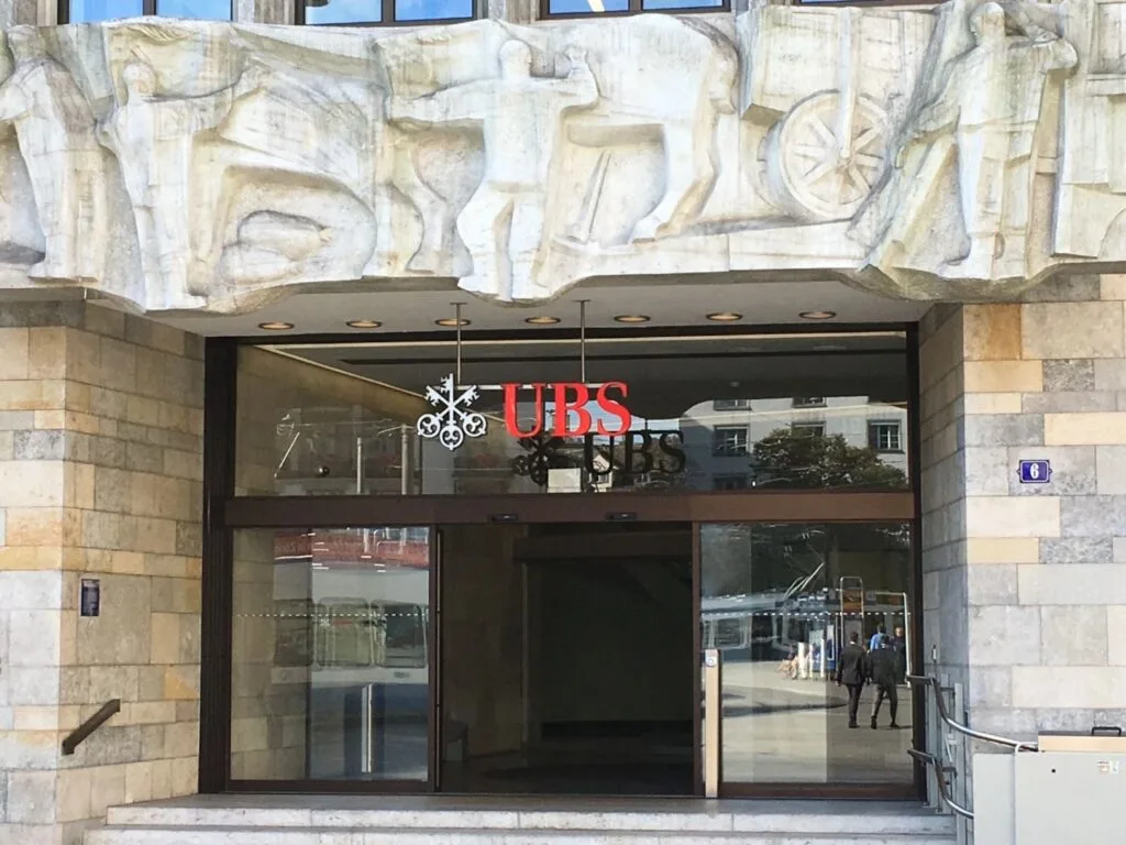 Entrance to the UBS bank branch at Paradeplatz 6 in Zurich, Switzerland, featuring a red UBS logo and a monumental stone relief sculpture by Franz Fischer above the glass doors.