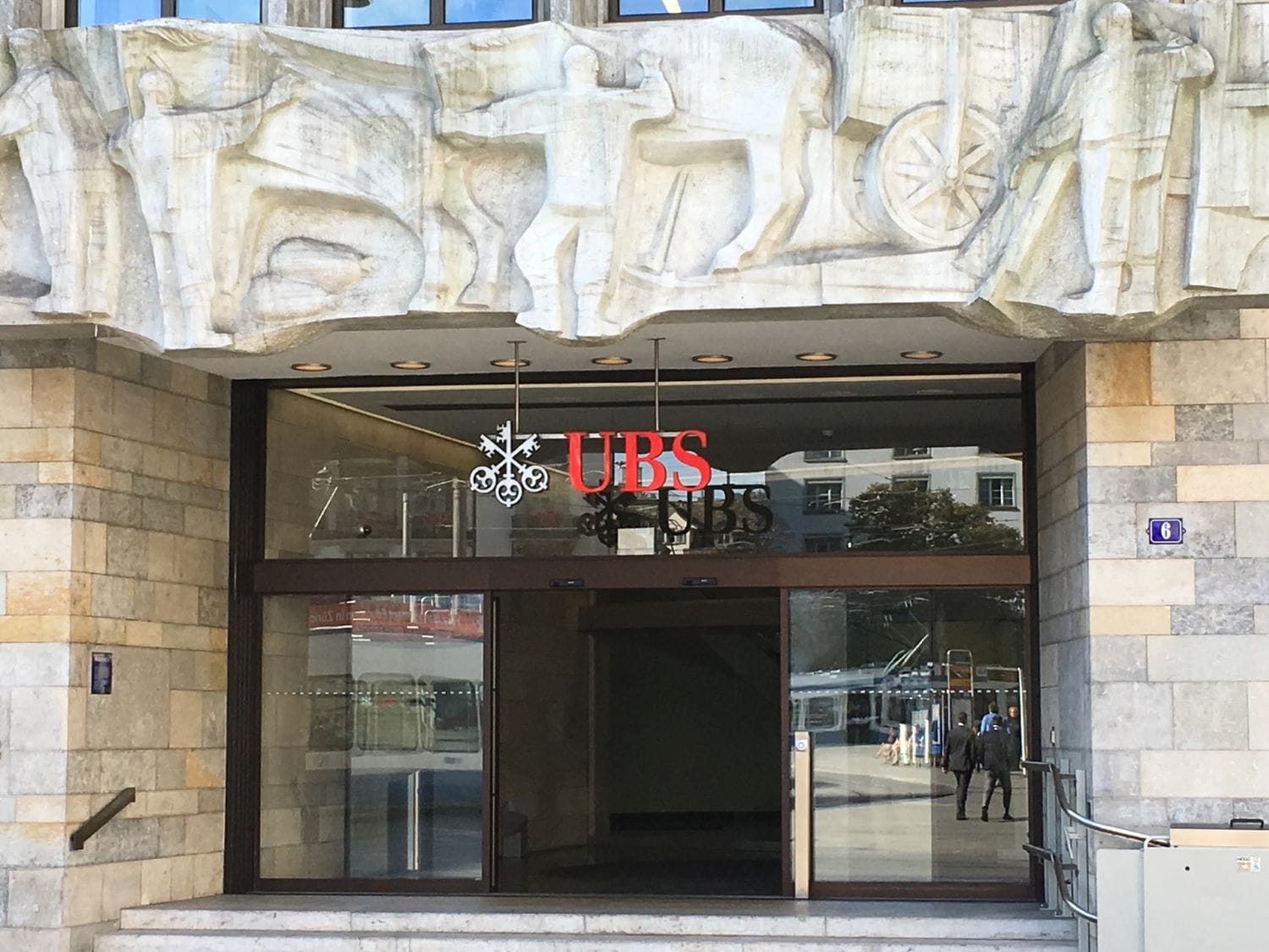 Entrance to the UBS bank branch at Paradeplatz 6 in Zurich, Switzerland, featuring a red UBS logo and a monumental stone relief sculpture by Franz Fischer above the glass doors.